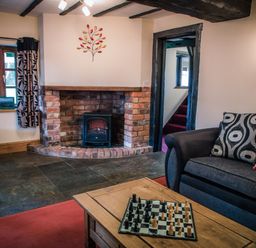 A living room with a brick fireplace and a wood stove a dark sofa with patterned cushions a wooden coffee table with a chessboard and a window with curtains at Brambles Cottage in Little Malvern near Malvern