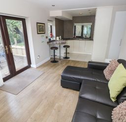 An open living room and kitchen area with a black leather sofa with cushions wooden floor a small bar with stools and double glass doors leading to an outdoor deck at The Garden Flat in Saundersfoot