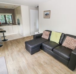 A living area with a black sectional sofa with three cushions and a small kitchen with bar stools at The Garden Flat in Saundersfoot