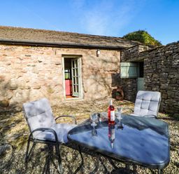An outdoor patio with a stone wall a glass table with two cushioned chairs a bottle of wine and two glasses at Cherry Tree Cottage Soulby near Kirkby Stephen