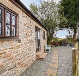 An outdoor seating area with gravel ground a stone building with windows and door benches and a table with chairs at Brocks Barn Sweetshouse near Lostwithiel