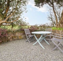An outdoor seating area with a round table and three chairs on gravel next to a stone wall and trees at Brocks Barn Sweetshouse near Lostwithiel