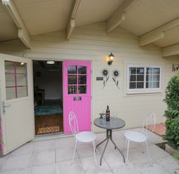 A patio with a round table and two chairs outside a cabin with pink and cream double doors at The Log Cabin in Bushton near Royal Wootton Bassett