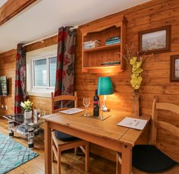 A wooden dining table with two chairs wine bottle and glasses next to a window with curtains and a small glass coffee table in a wooden room at The Log Cabin in Bushton near Royal Wootton Bassett