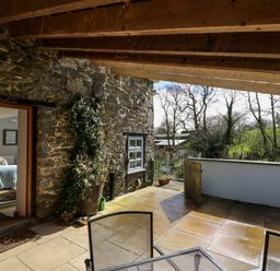 A stone patio with metal chairs and a wooden pergola next to a stone wall and an open door showing a living room at The Barn at Glanoer Hundred House near Builth Wells