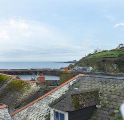 A view of rooftops with chimneys overlooking a harbor and green hillside at Thimble Cottage in Mevagissey
