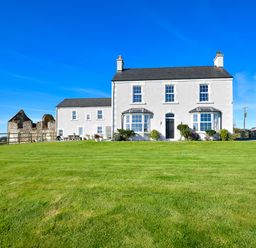A white two story house with bay windows and a green lawn under a blue sky at Ty Carmel in Rhosneigr