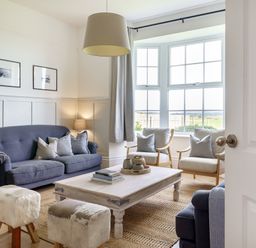 A living room with blue sofas wooden chairs a wooden coffee table and large windows at Ty Carmel in Rhosneigr