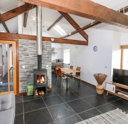 An open living room with exposed wooden beams a wood burning stove a dining table chairs a TV on a stand and a window at Y Beudy in Criccieth
