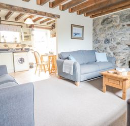 A living room with two gray sofas a wooden coffee table and a stone wall next to a kitchen and dining area at Ysgybor Yd in Chwilog near Criccieth