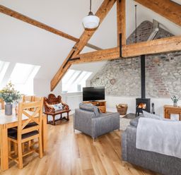 An open living and dining area with a wooden dining table and chairs grey sofas and a wood burning stove under exposed wooden beams at Yr Hen Festri in Y Felinheli