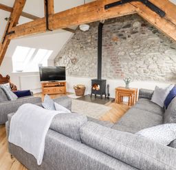 A living room with a gray sofa, armchair, wood burning stove, TV on a wooden stand, and exposed wooden beams at Yr Hen Festri in Y Felinheli