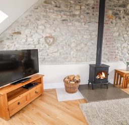 A living room with a stone wall a wood burning stove a basket of firewood a television on a wooden stand and nested wooden tables at Yr Hen Festri in Y Felinheli