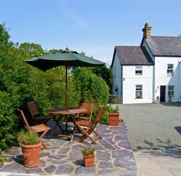 An outdoor patio with wooden table and chairs under a green umbrella near a white house with a gravel driveway at Carreg Rhys in Malltraeth near Newborough
