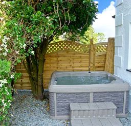 An outdoor hot tub next to a house with a small tree and wooden fence at Carreg Rhys in Malltraeth near Newborough