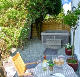 A small outdoor garden area with a hot tub, wooden fence, table with a checked tablecloth, bottle of wine, cake, chairs, and potted plants at Carreg Rhys in Malltraeth near Newborough