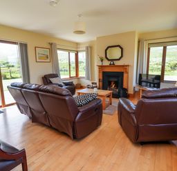 A living room with brown leather sofas a fireplace a wooden coffee table and large windows at Brandon Holiday Home in Cloghane County Kerry