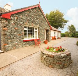A stone and white house with red trim a red bench and a circular stone flower bed on a gravel driveway at The Lodge Churchtown near Beaufort County Kerry
