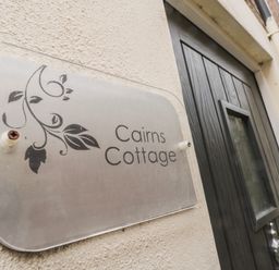 A sign with leaves and the text Cairns Cottage on a wall near a dark wooden door at Cairns Cottage in Whitby