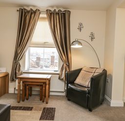 A living room with a black armchair pillow wooden tables window with curtains a floor lamp wooden TV stand and a bar stool at Cairns Cottage in Whitby