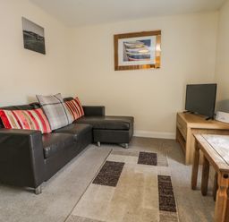 A living room with a black leather sofa with cushions a wooden table a television on a wooden stand and two pictures on the walls at Cairns Cottage in Whitby