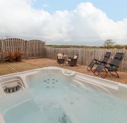 An outdoor area with a hot tub two black recliners a small table with two chairs and a wooden fence at Dairy Barn in Pershore