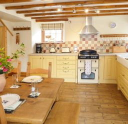 A kitchen with wooden beams yellow cabinets a wooden dining table with pink flowers at Fell View Cottage in Bootle near Millom