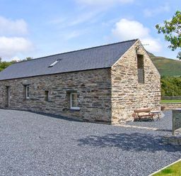 A stone barn with a slate roof and gravel driveway surrounded by trees and hills at Garth Morthin The Barn in Porthmadog