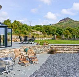 An outdoor patio area with metal and wooden chairs tables stone walls and green grass with a hill in the background at Garth Morthin The Barn in Porthmadog