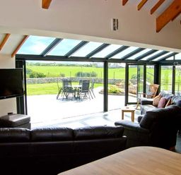 A living room with black leather sofas a wall-mounted TV and large glass doors opening to a patio with outdoor furniture at Garth Morthin The Barn in Porthmadog