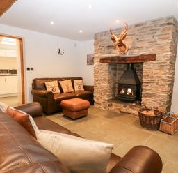 A living room with brown leather sofas a stone fireplace with a wood burner and a doorway leading to a kitchen at Garth Morthin The Stables in Porthmadog