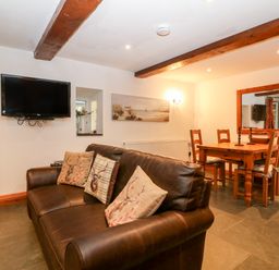 A living room with a brown leather sofa and cushions a wall mounted TV a wooden dining table with chairs and a large wall mirror at Garth Morthin The Stables in Porthmadog