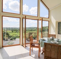 A dining area with a table and chairs next to large windows and doors opening to a garden with a view of fields at Llety'r Wennol in Ffarmers near Lampeter