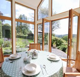 A dining area with a round table set with dishes and cutlery surrounded by wooden chairs and glass doors opening to a garden at Llety'r Wennol Ffarmers near Lampeter