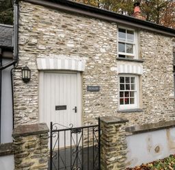 An exterior view of a stone cottage with a white door two windows and a black metal gate at Penlon in Capel Bangor near Aberystwyth