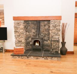 A stone fireplace with a wood burning stove burning fire a wooden box and fireplace tools on wooden floor at Fernleigh Cottage in Allithwaite near Grange over Sands