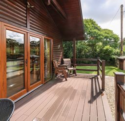 A wooden deck with two wooden chairs outside a house with large glass doors at Barn Shelley Lodge in Copplestone near Crediton
