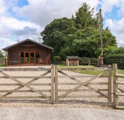 A wooden gate leading to a gravel driveway with a wooden cabin and trees in the background at Barn Shelley Lodge in Copplestone near Crediton