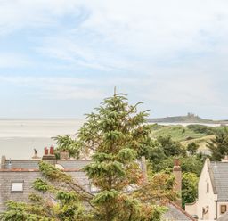 Rooftops of houses with trees in front and a beach with a castle on a hill in the distance at 3B Coastguard Cottages in Low Newton-By-The-Sea