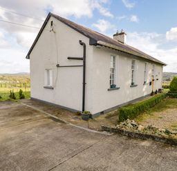 An exterior view of a white house with a sloped roof and multiple windows surrounded by a garden and concrete driveway at The Old School House in Gortermone near Carrigallen County Leitrim