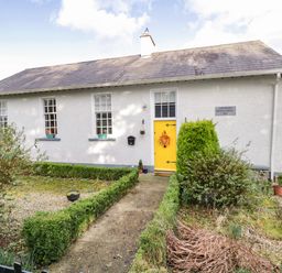 A single story white building with a yellow door and three windows surrounded by bushes and a garden path at The Old School House in Gortermone near Carrigallen County Leitrim