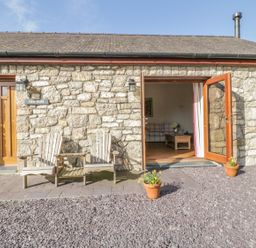 Exterior view of a stone building with wooden chairs outside and open door showing part of a living room at Hen Stabl Llangoed near Beaumaris