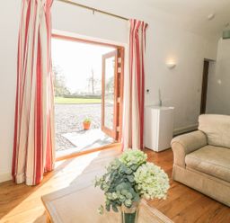 A living room with a sofa a coffee table with flowers wooden floors and open double doors with red and white striped curtains at Hen Stabl in Llangoed near Beaumaris