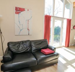 A black leather sofa with a red blanket folded on it next to tall windows with red curtains in a living area at Orchard Lodge in Warkworth