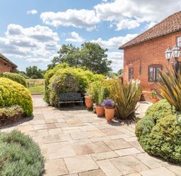 A patio with stone tiles surrounded by bushes and potted plants between two brick buildings at Horseshoe in Little Glemham near Wickham Market