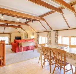 A dining area with a table and chairs next to a living room with red sofas and wooden beams on the ceiling at Hayloft Cottage in Little Glemham near Wickham Market
