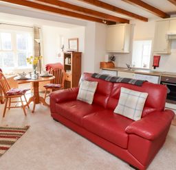 A living room with a red leather sofa two wooden chairs around a round wooden table and visible kitchen cabinets at The Bothy in Lowick near Holy Island