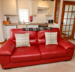 A red leather sofa with two plaid cushions in a living space with a small kitchen area and a wooden door at The Bothy Lowick near Holy Island