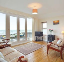 A living room with wooden armchairs a rug a television and large glass doors overlooking the sea at Trysor Y Mor in Borth