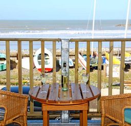 A wooden balcony with two wicker chairs and a wooden table with a bottle and two glasses overlooking boats on a beach at Trysor Y Mor in Borth